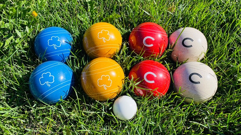 Op de afbeelding: Een set jeu de boules ballen in groen gras. De set bevat blauwe ballen met witte klavers, gele ballen met witte strepen en klavers, rode ballen met een witte "C" en witte ballen met een zwarte "C".