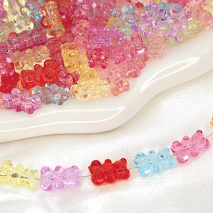 May include: A collection of colorful gummy bear-shaped beads in various shades of pink, red, yellow, and blue. Some beads are scattered on a white dish, while others are strung on a wire, creating a necklace.