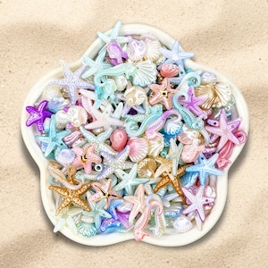 May include: A white, flower-shaped dish filled with an assortment of colorful, miniature sea creatures. The collection includes starfish, seashells, and seahorses in shades of pink, blue, gold, and purple. The dish sits on a sandy surface.