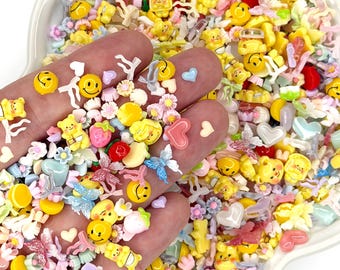 May include: A hand holding a variety of colorful resin embellishments, including yellow smiley faces, teddy bears, flowers, hearts, and butterflies. The embellishments are in a white, flower-shaped dish, and are ideal for crafting and scrapbooking projects.