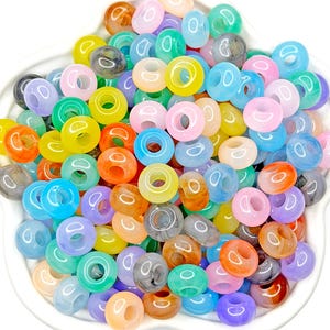 May include: A collection of round, smooth, translucent and opaque plastic beads in a wide variety of vibrant colors including pink, blue, green, yellow, orange, and grey. The beads feature a central hole and are piled together in a cluster.