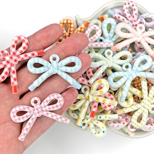 May include: A collection of colorful bow-shaped hair accessories in various patterns, including gingham and polka dots. The bows are in shades of red, pink, blue, and yellow. Some bows are held in a hand, while others are in a white dish.