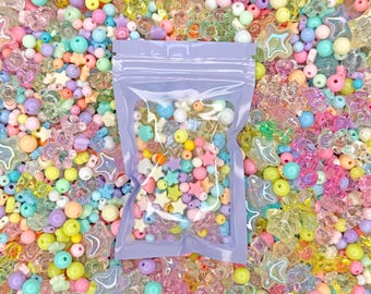 May include: A clear plastic bag filled with colorful beads in various shapes and sizes, including stars, circles, and bears. The beads are in shades of pink, yellow, blue, and green. The bag is set against a background of similar beads.