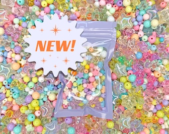Puede incluir: Una bolsa de plástico transparente llena de cuentas de colores, estrellas y otras formas, con la palabra "NEW!" en una pegatina blanca en forma de estrella. Las cuentas son de varios colores pastel.