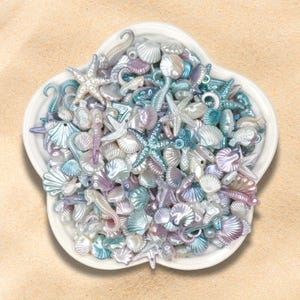 May include: A white, flower-shaped dish filled with an assortment of iridescent seashell beads. The beads include starfish, seahorses, and various shell shapes in shades of blue, purple, and white. The dish sits on a sandy surface.