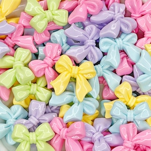May include: A collection of pastel-colored bows in shades of pink, yellow, blue, green, and purple. The bows are glossy and appear to be made of resin or a similar material. The bows are scattered, creating a colorful and playful arrangement.