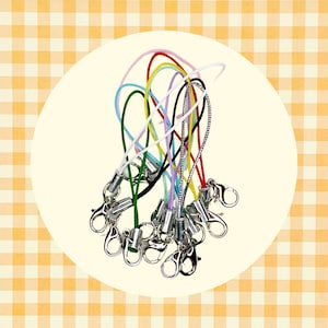 May include: A collection of colorful lanyards with metal lobster claw clasps. The lanyards feature a variety of colors, including red, green, blue, yellow, and black. The clasps are silver-toned and appear to be made of metal. The background is a yellow and white checkered pattern.