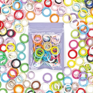 May include: A collection of small, circular metal spring gate rings in a variety of colors including red, blue, green, yellow, pink, orange, and black. A portion of these rings is contained within a clear plastic bag with a purple top.