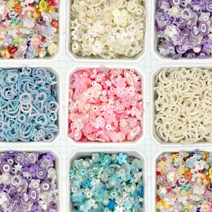 May include: An overhead shot of nine square containers filled with various colorful beads. The beads include floral, circular, and star shapes in shades of pink, blue, purple, yellow, and white. The containers are arranged in a grid pattern.