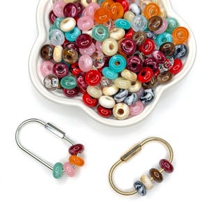 May include: A white flower-shaped bowl filled with a variety of colorful, translucent, and opaque round beads. Below the bowl are two oval-shaped metal carabiner clips, one silver and one gold, each featuring a few beads threaded onto the metal loop.