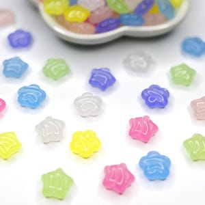 May include: A collection of colorful, translucent star-shaped beads in various shades of blue, green, pink, yellow, and white. Some beads are scattered on a white surface, while others are in a white, petal-shaped dish. The beads are small and appear to be made of plastic or resin.
