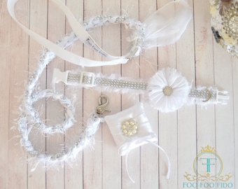 May include: White dog collar and leash set with a white flower charm and a small white pouch with a pearl embellishment. The collar and leash are adorned with rhinestones and white fur. The text "Foo Foo Fido" is printed on the leash.