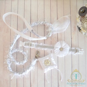 May include: White dog collar and leash set with a white flower charm and a small white pouch with a pearl embellishment. The collar and leash are adorned with rhinestones and white fur. The text "Foo Foo Fido" is printed on the leash.