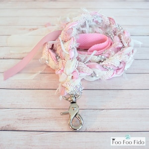 May include: Pink and white braided dog leash with a silver clip. The leash is made of fabric and ribbon and has a delicate, feminine look.