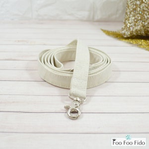 May include: A white cotton dog leash with a silver clip. The leash is coiled up on a light wood surface.