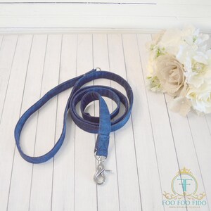 May include: A blue denim dog leash with a silver clip. The leash is coiled on a white wooden surface.