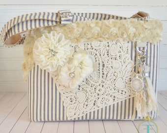 May include: A white and blue striped fabric tote bag with a white lace overlay, white fabric flowers, and a white fluffy trim. The bag has a silver key chain with a charm attached.