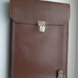 May include: A brown leather portfolio with a flap closure and a silver-colored clasp. The portfolio has a rectangular shape and a small leather strap with a button closure at the bottom. The leather has a slightly worn appearance.