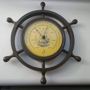 May include: A dark brown ship wheel barometer with a yellow circular face. The dial features a central illustration of a sailing ship, black numerical markings, and Cyrillic text including "НОРМАЛЬНОЕ", "НИЗКОЕ", and "ВЫСОКОЕ" with two black needles.