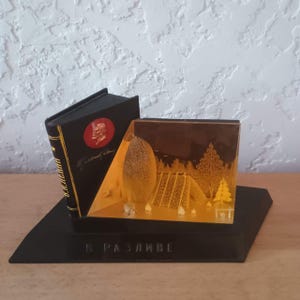 May include: A black desk ornament featuring a miniature book with a red circular emblem and gold Cyrillic text. Attached is a transparent amber-colored block displaying a relief scene of a forest, a small hut, and trees. The base bears the text В РАЗЛИВЕ.
