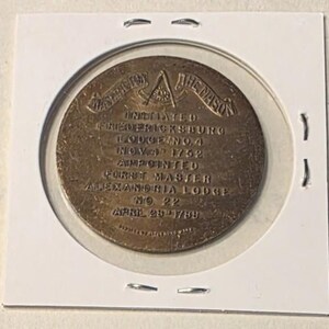 Puede incluir: Medalla mas&oacute;nica de color bronce, dentro de un soporte de pl&aacute;stico transparente. La medalla presenta texto que incluye "WASHINGTON THE MASON" y "FREDERICKSBURG LODGE NO. 4 NOV. 4 1752". La medalla redonda tiene un dise&ntilde;o detallado.