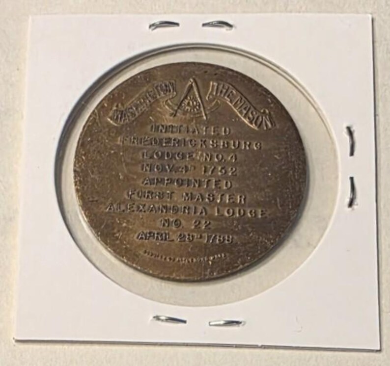 Puede incluir: Medalla mas&oacute;nica de color bronce, dentro de un soporte de pl&aacute;stico transparente. La medalla presenta texto que incluye "WASHINGTON THE MASON" y "FREDERICKSBURG LODGE NO. 4 NOV. 4 1752". La medalla redonda tiene un dise&ntilde;o detallado.