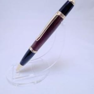 May include: A handcrafted pen with a dark brown wooden barrel, black accents, and gold-colored metal trim. The pen is displayed on a clear acrylic stand, showcasing its elegant design. The pen's tip and top are also gold-colored.