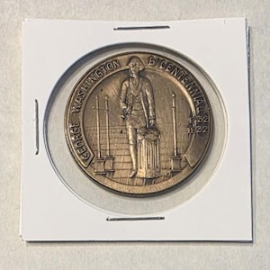 Puede incluir: Una moneda conmemorativa de color bronce que representa a George Washington. La moneda est&aacute; inscrita con "GEORGE WASHINGTON BICENTENNIAL 1932". La imagen muestra a Washington de pie junto a un pilar.