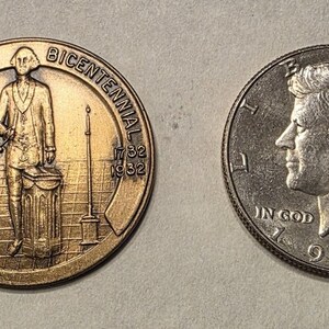 Puede incluir: Dos monedas: una medalla conmemorativa de bronce con George Washington y el texto "George Washington Bicentennial 1732 1932", y una moneda de medio d&oacute;lar Kennedy de plata de 1986 con el texto "In God We Trust".