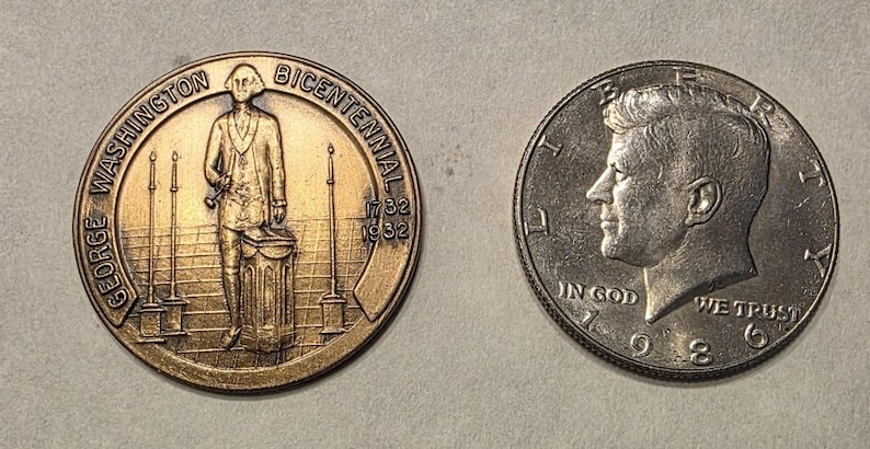 Puede incluir: Dos monedas: una medalla conmemorativa de bronce con George Washington y el texto "George Washington Bicentennial 1732 1932", y una moneda de medio d&oacute;lar Kennedy de plata de 1986 con el texto "In God We Trust".