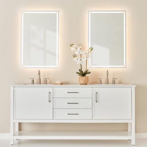 Puede incluir: Un mueble de tocador doble blanco con encimera de mármol, dos espejos rectangulares con bordes iluminados y una planta decorativa. El mueble tiene dos puertas, tres cajones y una balda inferior.