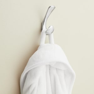 May include: A shiny chrome hook holds a fluffy white hooded bathrobe. The robe is made of a soft, plush material. The hook is mounted on a light beige wall, creating a clean and simple aesthetic. The robe's hood has small, rounded ears.