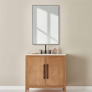 Könnte beinhalten: Ein Badezimmerwaschtisch aus hellem Holz mit weißer Arbeitsplatte und schwarzer Hardware. Über dem Waschtisch hängt ein rechteckiger Spiegel mit schwarzem Rahmen. Ein schwarzer Wasserhahn und eine kleine dekorative Flasche befinden sich auf der Arbeitsplatte.