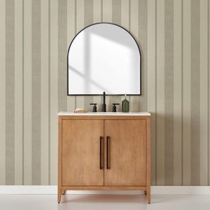 Könnte beinhalten: Ein Badezimmer-Waschtisch aus hellem Holz mit einer weißen Arbeitsplatte, einem schwarzen Wasserhahn und einem schwarz gerahmten, bogenförmigen Spiegel. Der Waschtisch hat zwei Türen mit dunklen Griffen. Eine Seifenschale und eine grüne Flasche befinden sich auf der Arbeitsplatte. Der Hintergrund ist eine gestreifte Tapete.