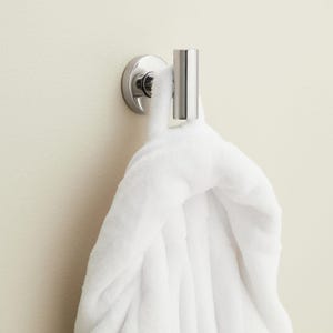 May include: A chrome-finished wall hook with a cylindrical post and a circular base. A fluffy white robe is hanging from the hook. The hook is mounted on a light-colored wall, creating a clean and minimalist aesthetic.