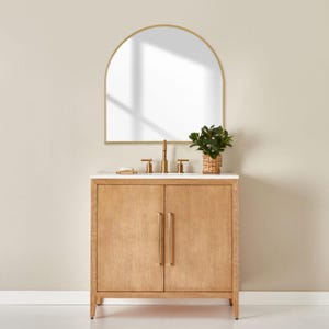 Könnte beinhalten: Ein helles Holzwaschbecken mit weißer Arbeitsplatte und goldenen Armaturen. Über dem Waschtisch befindet sich ein goldgerahmter, bogenförmiger Spiegel. Eine kleine Pflanze in einem geflochtenen Korb steht auf der Arbeitsplatte. Der Waschtisch hat zwei Türen mit langen, vertikalen goldenen Griffen.