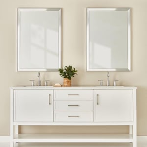 May include: A white bathroom vanity with a rectangular countertop, two mirrors, and a small plant in a woven basket. The vanity has two cabinets and three drawers with silver hardware. The mirrors have silver frames.