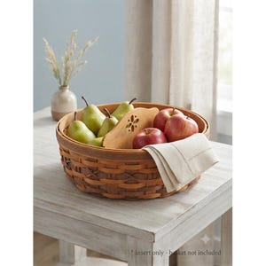 May include: A round woven wooden basket with a central handle featuring a cutout flower design. The basket contains green pears and red apples, with a folded cream-colored linen napkin draped over the side, showcasing a subtle stitched border.