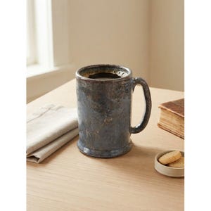 Peut inclure: Une tasse en céramique cylindrique avec un vernis moucheté bleu foncé et brun, présentant une finition texturée et rustique. La tasse possède une anse incurvée robuste sur le côté et une base légèrement évasée, offrant un aspect profond et patiné.
