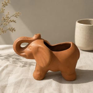 Op de afbeelding: Een terracotta olifantvormige plantenbak met een grote opening op de rug. De plantenbak heeft een omhoog gekrulde slurf en vier stevige poten. De olifant heeft een warme, aardse tint, perfect voor kleine planten.