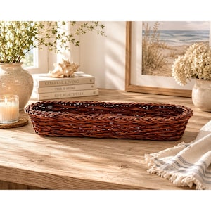 Puede incluir: Una cesta larga y rectangular de mimbre tejido en color marrón oscuro con un borde trenzado. La cesta presenta un diseño robusto de parte superior abierta con un patrón de tejido apretado y consistente en toda su estructura funcional.