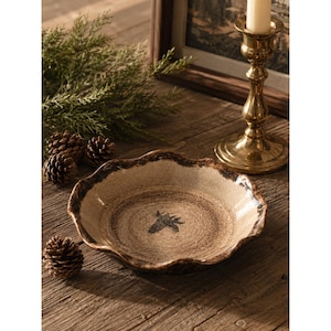 May include: A round ceramic bowl with a scalloped edge, featuring a speckled beige and brown glaze. The center of the bowl displays a small, dark, hand-drawn illustration of a pinecone. The bowl has a rustic, textured finish with darker brown tones along the rim.