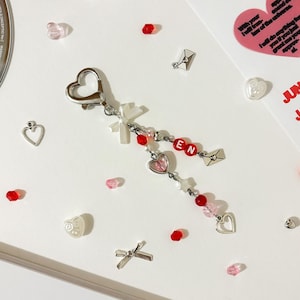 Puede incluir: Un llavero plateado con un cierre en forma de corazón y las letras "EN" en cuentas rojas. El llavero está adornado con dijes de corazón y estrella, cuentas rojas y rosas, y un pequeño lazo. Hay pegatinas en forma de corazón y otros elementos decorativos dispersos.