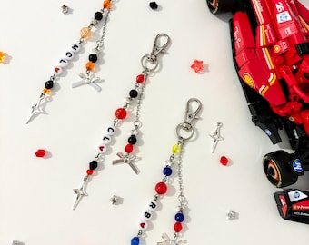 May include: Three beaded keychains with silver clasps and star charms. One keychain spells out "MCL" with black, white, and orange beads. Another spells "RB" with red, blue, and yellow beads. A red race car toy is in the background.