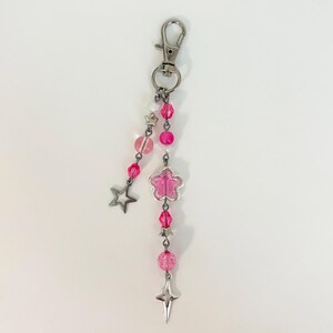 May include: A pink and silver keychain with a lobster clasp. The keychain features pink beads, a flower-shaped charm, and star-shaped charms. The beads are various shades of pink, and the charms are silver.