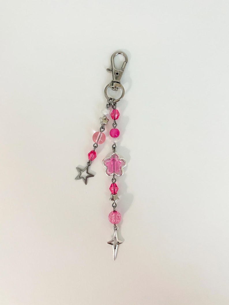 May include: A pink and silver keychain with a lobster clasp. The keychain features pink beads, a flower-shaped charm, and star-shaped charms. The beads are various shades of pink, and the charms are silver.