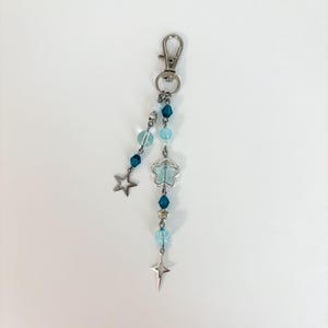 May include: A silver-toned keychain with a lobster clasp, adorned with blue and clear beads, a star charm, and a flower-shaped charm. The keychain is designed with a celestial theme, featuring a star and a cross-shaped charm.