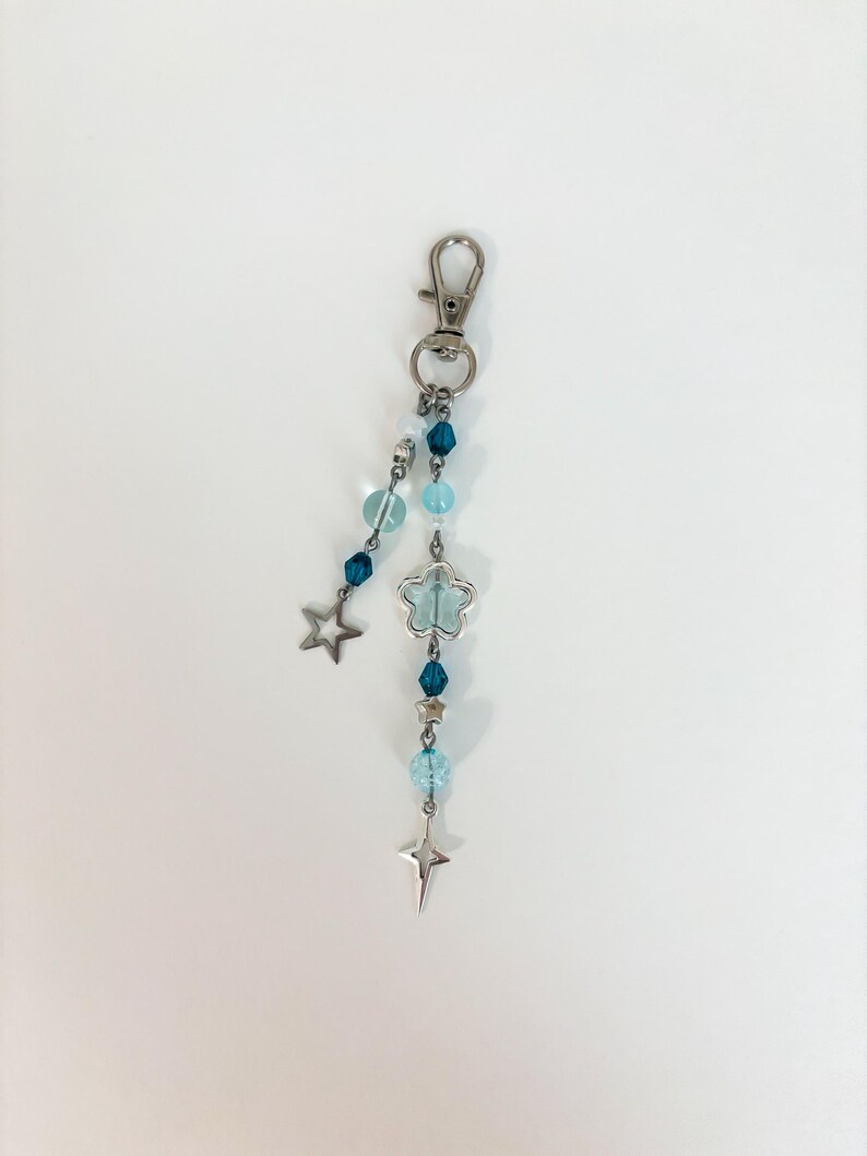 May include: A silver-toned keychain with a lobster clasp, adorned with blue and clear beads, a star charm, and a flower-shaped charm. The keychain is designed with a celestial theme, featuring a star and a cross-shaped charm.