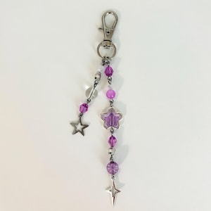May include: A silver-toned keychain with a lobster clasp. The keychain features a dangling design of purple beads, a clear bead, a purple flower charm, and two star charms. The beads are various shapes and sizes.