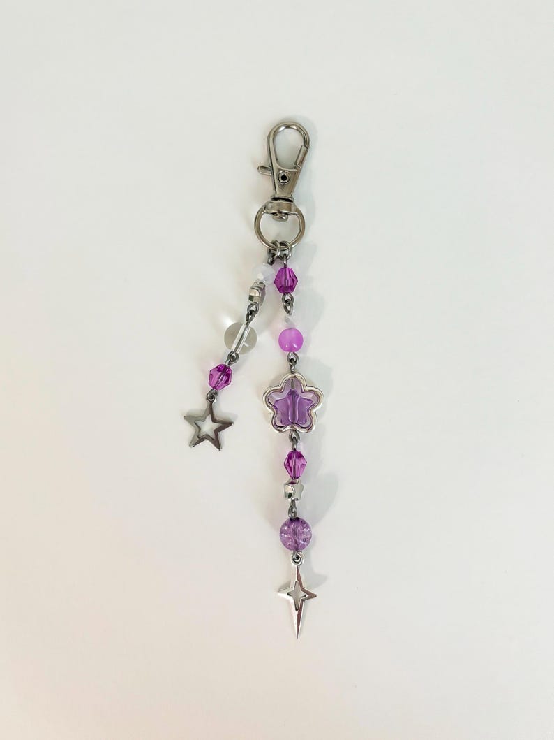 May include: A silver-toned keychain with a lobster clasp. The keychain features a dangling design of purple beads, a clear bead, a purple flower charm, and two star charms. The beads are various shapes and sizes.